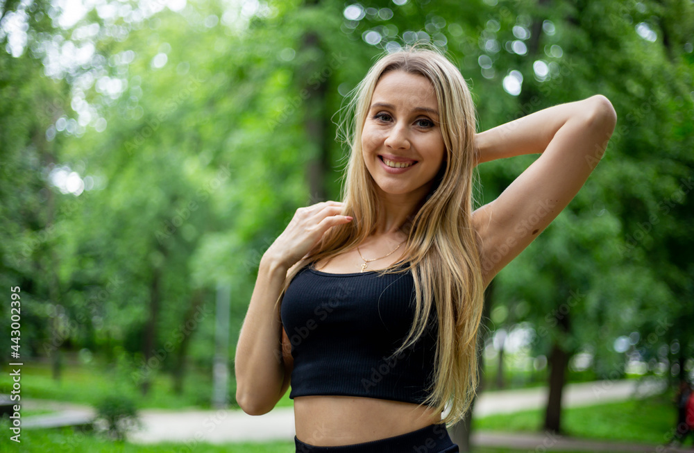 Young beautiful sporty blonde woman in a black T-shirt and in black tight sports shorts in good shape posing in front of green trees