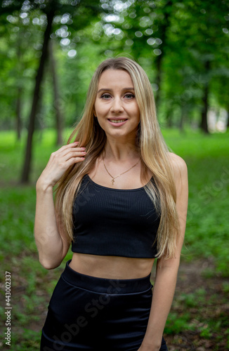 Young beautiful sporty blonde woman in a black T-shirt and in black tight sports shorts in good shape posing in front of green trees