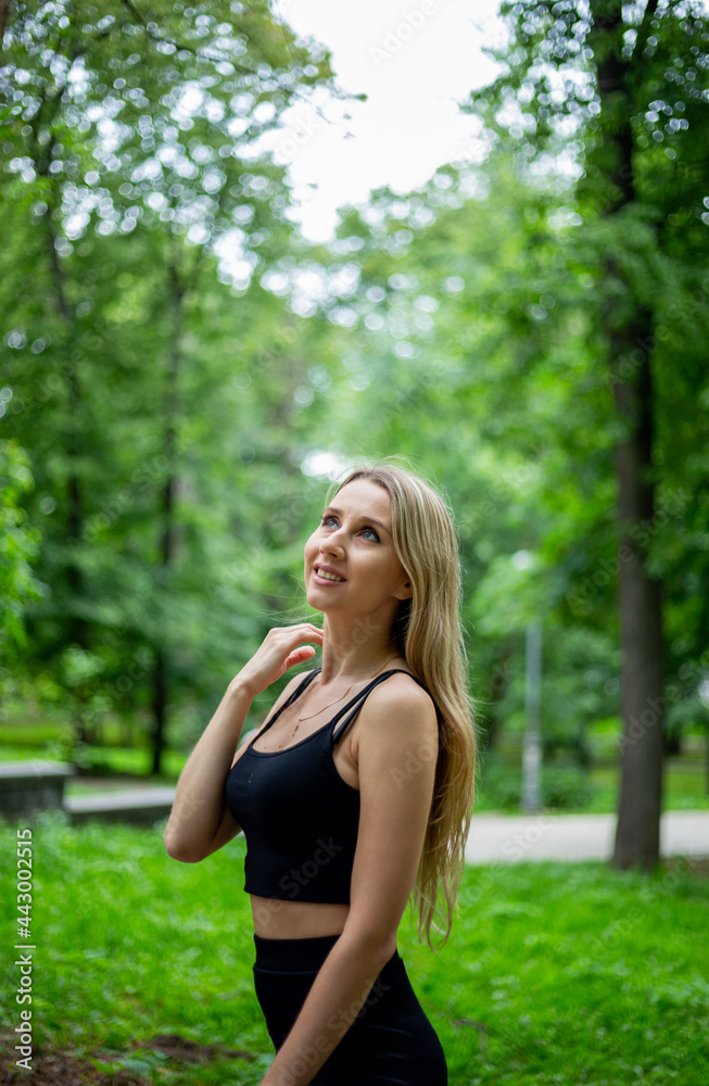 Young beautiful sporty blonde woman in a black T-shirt and in black tight sports shorts in good shape posing in front of green trees