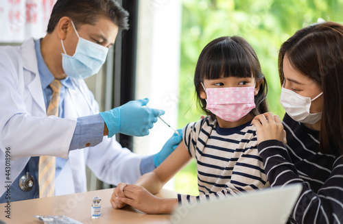 Asian senior doctor wearing gloves and isolation mask is making a COVID-19 vaccination in the shoulder of child patient with her mother at hospital.