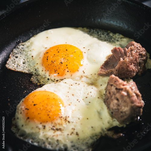 Men's breakfast - Two eggs are fried in a cast iron skillet with chunks of stew meat. Close-up