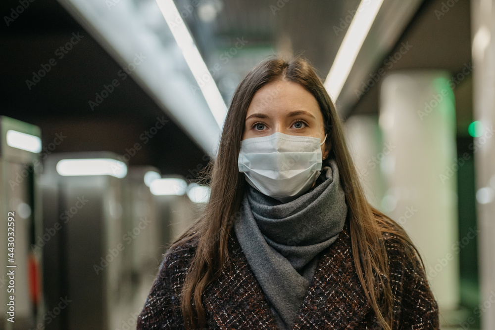 A close photo of a woman in a medical face mask to avoid the spread of coronavirus who is waiting for a train on the subway platform. Girl in a mask is keeping social distance in the underground.