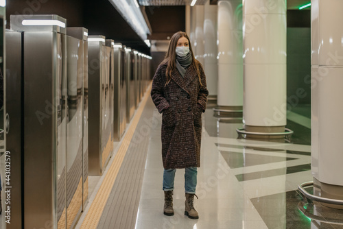 A woman in a medical face mask to avoid the spread of coronavirus is walking while waiting for a train on the subway platform. Girl in a surgical mask is keeping social distance in the underground.
