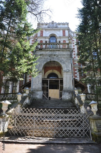 The overgrown and abandoned facade of the manor house of a noble estate made of red bricks with white trim. The main house of the Bykovo estate.  Wooden fence.
