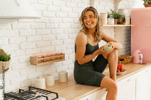 Laughing blond woman holding apple and posing on light kitchen. Healthy food concept.