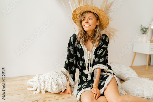 Graceful woman in bohemian dress sitting on pillows at home. Wearing earrings with feather and bracelets. White background in studio with pampas grass decor.