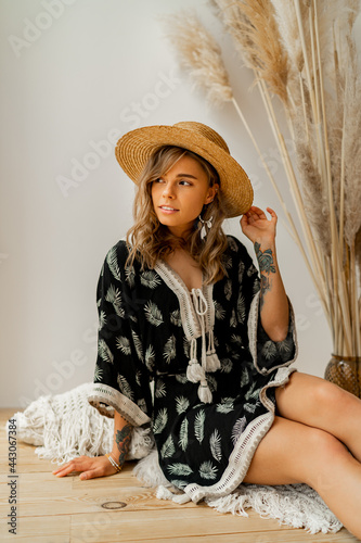 Pretty blond woman in straw hat and boho dress posing in studio over white background with pampas grass decor. Pillows with macrame ornament. Summer trends.