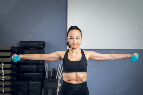 Front view of an Asian woman performing a side lateral raise exercise with two blue weights. Shoulder workout. I