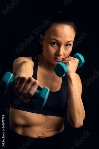 Vertical portrait, in studio, in front of a black background, of an Asian fighting athlete, Korean, boxing with weights in her hand, looking at the camera, with a serious, determined expression.