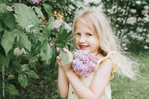 portrait of a thoughtful and sad little blonde girl in a yellow dress standing, smelling flowers near a blooming lilac in spring, the concept of a happy childhood and school holidays