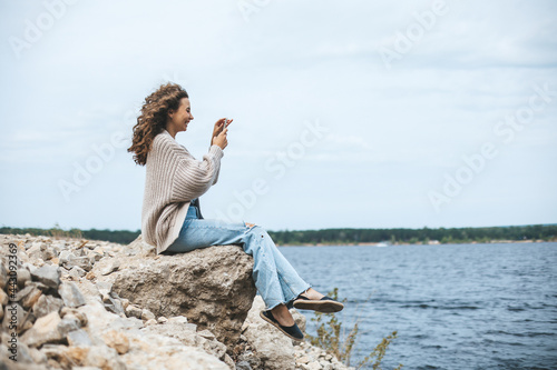 Young woman sitting on rocky bank taking photo of landscape using smartphone. photo