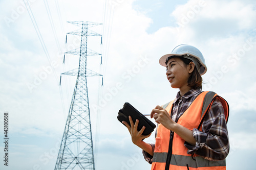 Electrical Asian beautiful engineer with high voltage electricity pylon and using walkie talkie and tablet to control assistant. Electrical power lines and towers at blue sky.