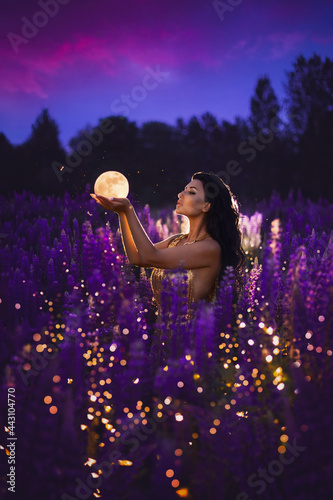 A brunette girl in a golden dress holding a moon in her hands and standing among a blooming purple lupine field with lights and fireflies. A magical night portrait.
