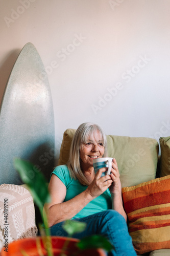 Smiling mature elder 60s woman sitting relaxing with cup of tea, coffee. Senior mid age stylish look woman portrait with cup looking away at surf style home photo