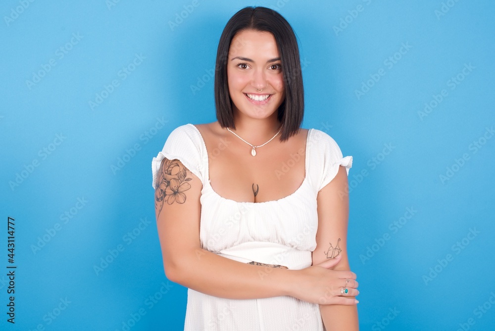 young beautiful tattooed girl wearing white dress standing against blue background laughing. young beautiful tattooed girl wearing white dress standing against blue background laughing.