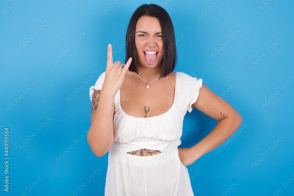 Portrait of a crazy young beautiful tattooed girl wearing white dress standing against blue background showing tongue horns up gesture, expressing excitement of being on concert of band. Portrait of a crazy young beautiful tattooed girl wearing white dress standing against blue background showing tongue horns up gesture, expressing excitement of being on concert of band.