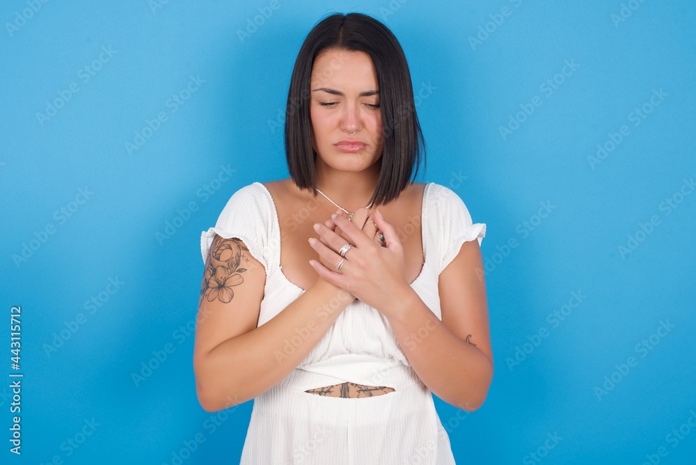 Sad young beautiful tattooed girl wearing white dress standing against blue background feeling upset while spending time at home alone staring at camera with unhappy or regretful look. Sad young beautiful tattooed girl wearing white dress standing against blue background feeling upset while spending time at home alone staring at camera with unhappy or regretful look.