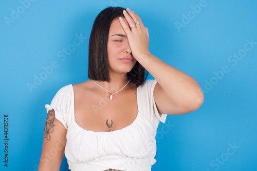 Frustrated young beautiful tattooed girl wearing white dress standing against blue background holding hand on forehead being depressed regretting what he did having headache, looking stressful.
