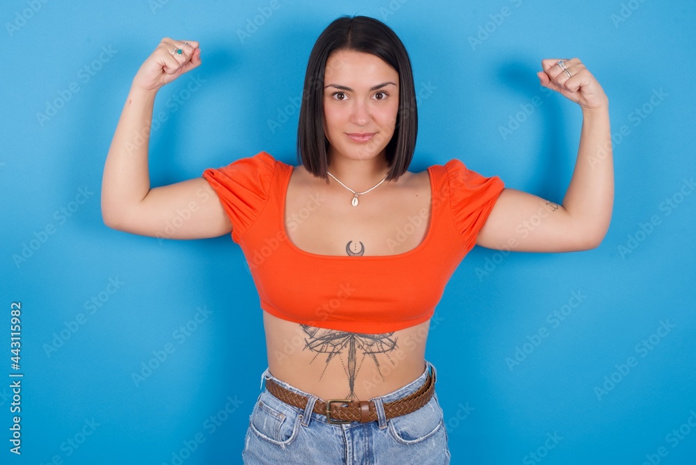 Waist up shot of young beautiful tattooed girl wearing orange crop top raises arms to show muscles feels confident in victory, looks strong and independent, smiles positively at camera. Sport concept. Waist up shot of young beautiful tattooed girl wearing orange crop top raises arms to show muscles feels confident in victory, looks strong and independent, smiles positively at camera. Sport concept.