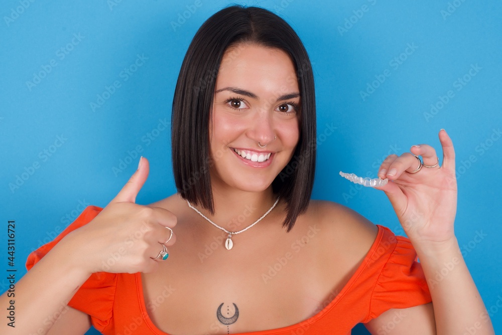 young beautiful tattooed girl wearing orange crop top standing against blue wall holding an invisible braces aligner and rising thumb up, recommending this new treatment. Dental healthcare concept. young beautiful tattooed girl wearing orange crop top standing against blue wall holding an invisible braces aligner and rising thumb up, recommending this new treatment. Dental healthcare concept.