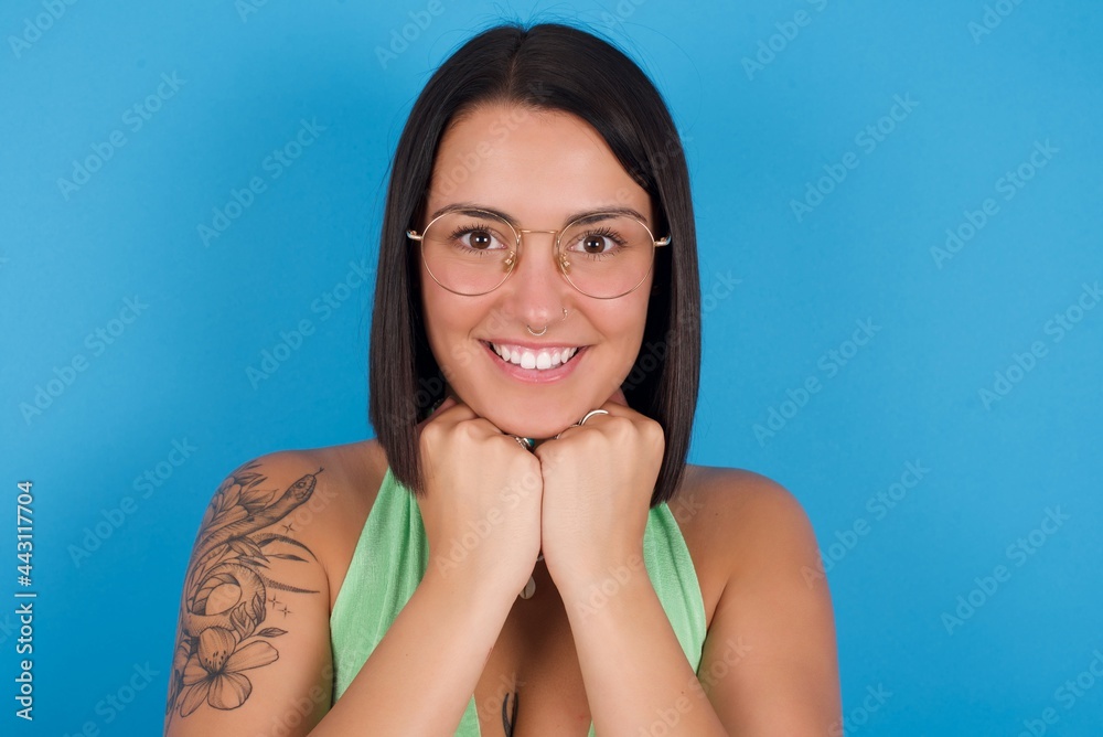 Satisfied young beautiful tattooed girl wearing green top standing against blue background touches chin with both hands, smiles pleasantly, rejoices good day with lover
