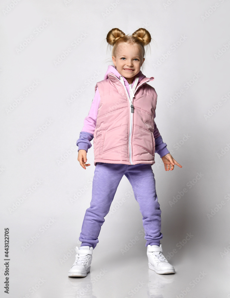 Positive cute blonde girl with a bagel hairstyle in a comfortable warm puffy vest with a hood, posing on a light background. Trendy kids fashion Positive cute blonde girl with a bagel hairstyle in a comfortable warm puffy vest with a hood, posing on a light background. Trendy kids fashion