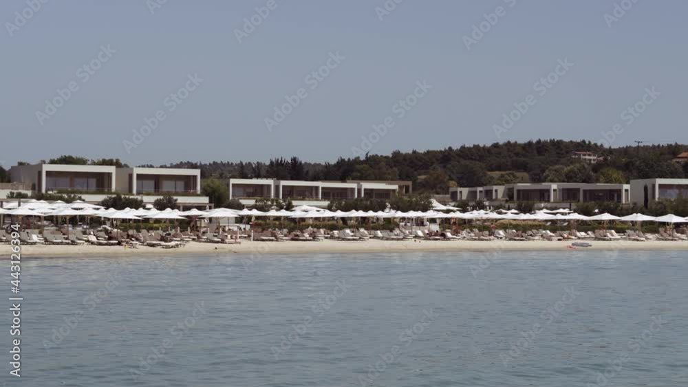 Modern seafront villas landscape with beach bathers on sea beds with umbrellas. Day view of Sani luxury resort in Greece at Kassandra Chalkidiki peninsula surrounded by green vegetation. Modern seafront villas landscape with beach bathers on sea beds with umbrellas. Day view of Sani luxury resort in Greece at Kassandra Chalkidiki peninsula surrounded by green vegetation.