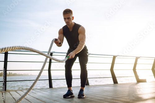 Young athlete wearing sports clothes is doing workout at the beach pier. Man doing exercises outdoors in the morning. Battle ropes. Kettlebell. Sport, Active life. 
