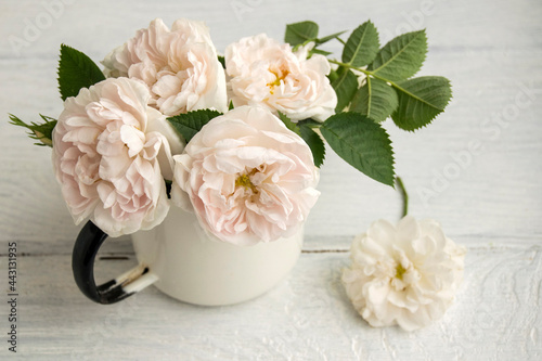 Delicate pink roses are in the old cup. The cup is on a white wooden table.