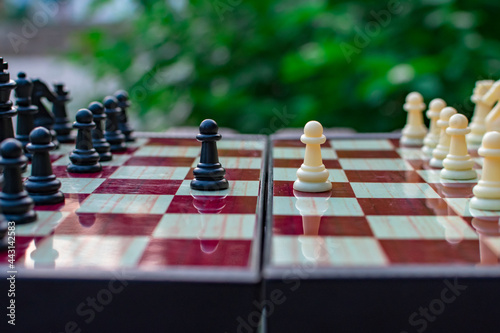 Chess pieces are placed on a chess board against a blurred background of nature. The pawns are black and white opposite each other. Blurred focus. Defocus. Shechamat day.