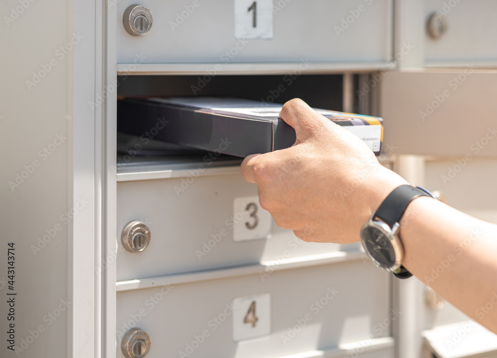 Mail man delivering small parcel in mailbox by house number, selective focus. Person’s hand holding package put in metal storage. People get packet from opened cabinet door. Sender, receiver concept. Mail man delivering small parcel in mailbox by house number, selective focus. Person’s hand holding package put in metal storage. People get packet from opened cabinet door. Sender, receiver concept.