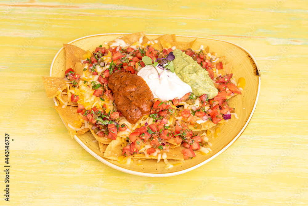 Yellow tray overflowing with Mexican corn nachos with guacamole, cream cheese, chili con carne and melted cheddar cheese on yellow table Yellow tray overflowing with Mexican corn nachos with guacamole, cream cheese, chili con carne and melted cheddar cheese on yellow table