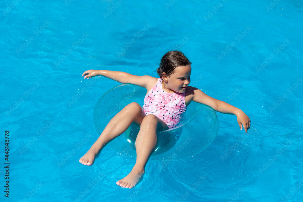 a happy smiling girl in a pink swimsuit floating on a blue transparent inflatable circle in the clear water of the sea on summer family holidays, active weekend