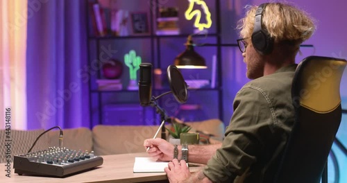 Portrait view of the caucasian man wearing glasses and headphones writing something while recording podcast at the studio with microphone. Working at the radio concept photo