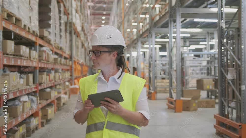 Woman manager or storekeeper works at retail or wholesale warehouse using tablet computer for inventory control walks between rows of racks, examining shelves with goods. Woman manager or storekeeper works at retail or wholesale warehouse using tablet computer for inventory control walks between rows of racks, examining shelves with goods.