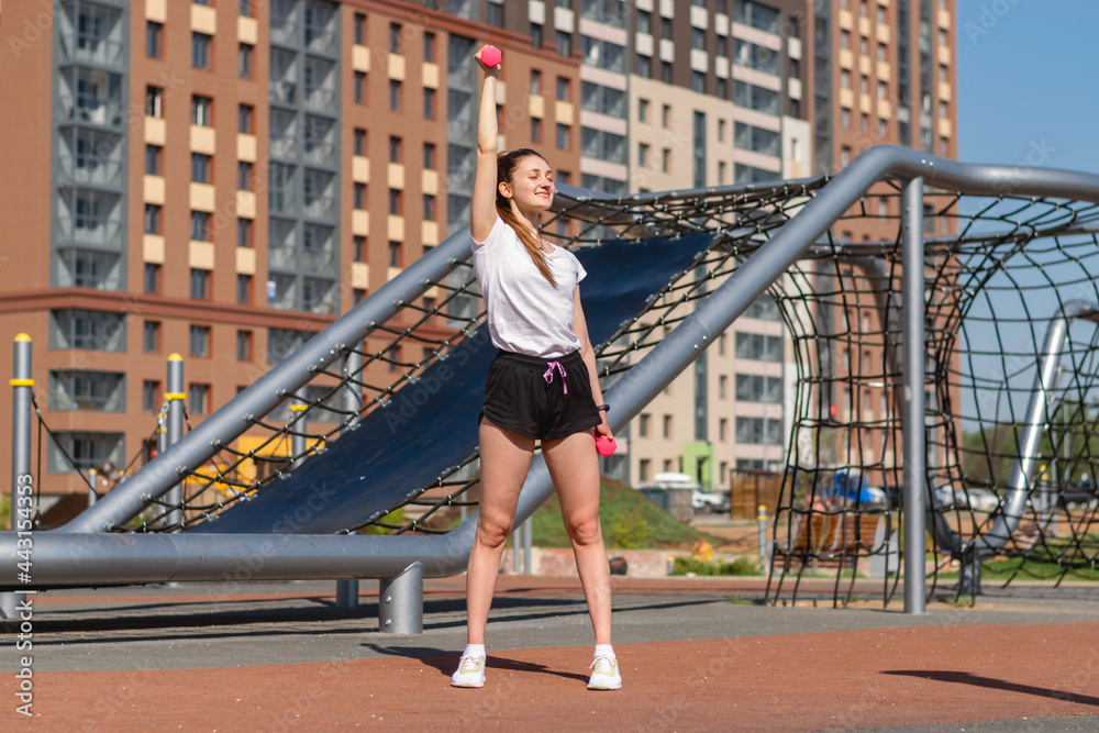 Young athletic woman in a white T-shirt,black shorts and white sneakers going in for sports,.doing a sports exercise with dumbbells on a street sports field.Sports healthy lifestyle.Outdoor training.