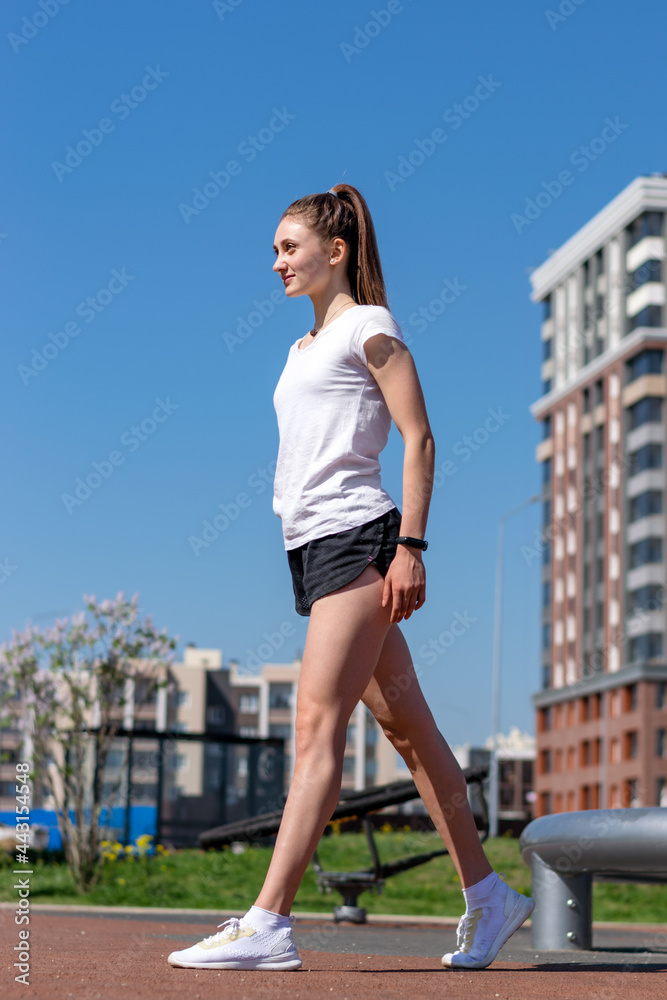 Young athletic woman in a white T-shirt,black shorts and white sneakers in full growth on a street sports field.Sports healthy lifestyle.Outdoor training.