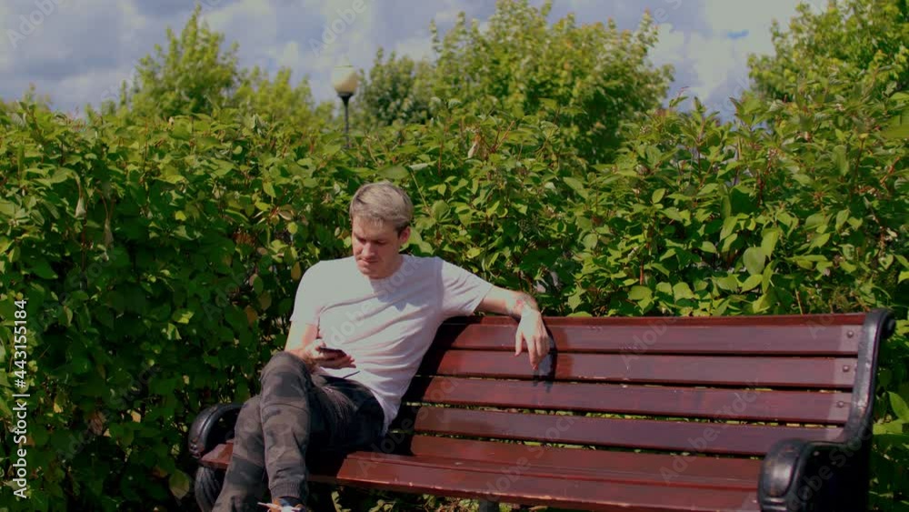 Young man with mobile phone rests, sitting on park bench surrounded by bushes. Guy speaking on smartphone, sitting on bench in parkland Young man with mobile phone rests, sitting on park bench surrounded by bushes. Guy speaking on smartphone, sitting on bench in parkland