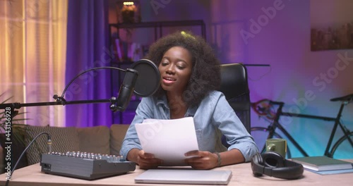 Portrait view of the multiracial young woman making a podcast audio recording at home at her studio while reading text from the paper. Shot on cinema camera in 4K photo