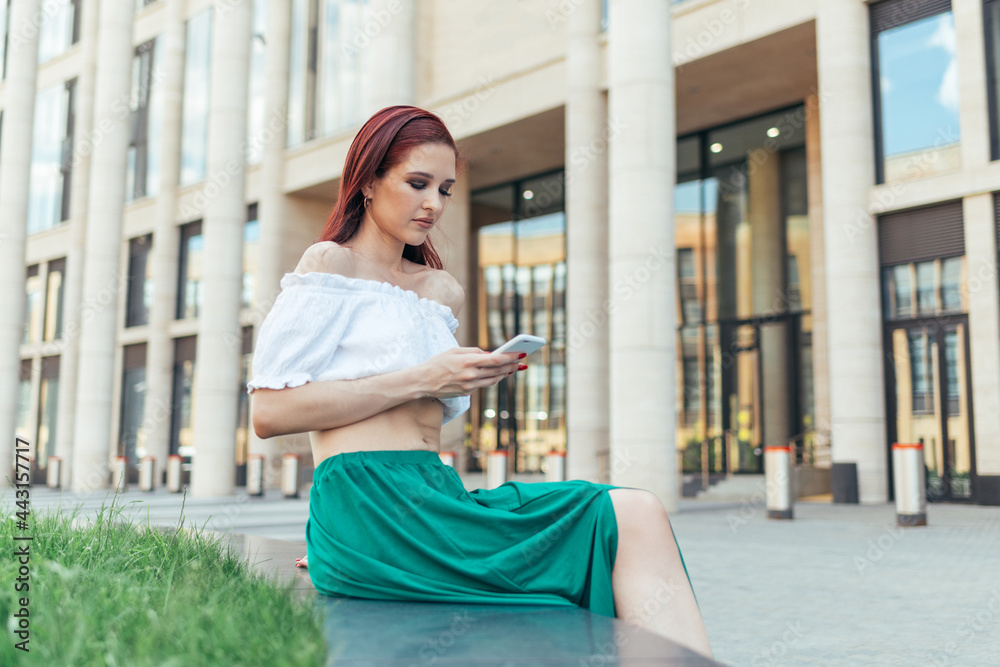 Redheaded woman in summer clothes sits on a bench and looks at her smartphone in front of a modern building