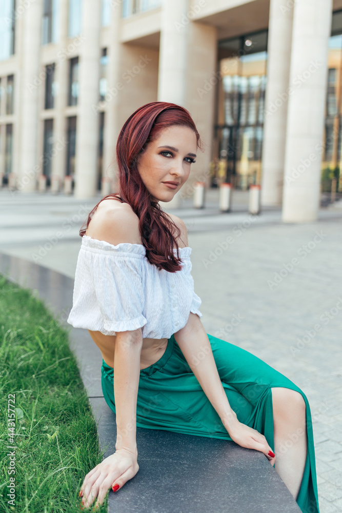 Red-haired woman in summer clothes sits on a bench against the background of a modern building