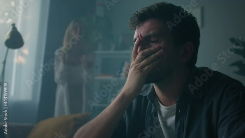 Irritated emotional woman shouting at man in living room at home, wife blaming and screaming at her husband holding his head, feeling anger and stress, bad mood. Scandals and quarrels in family.