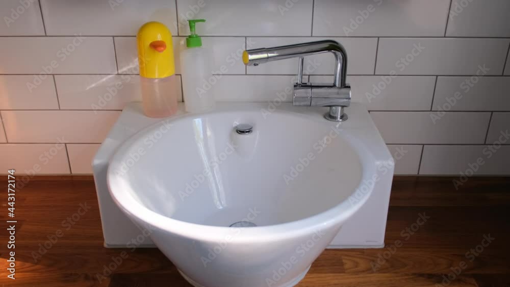 White sink with designed silver tape and different soaps in plastic bottles by white tile wall over wooden floor in bathroom White sink with designed silver tape and different soaps in plastic bottles by white tile wall over wooden floor in bathroom