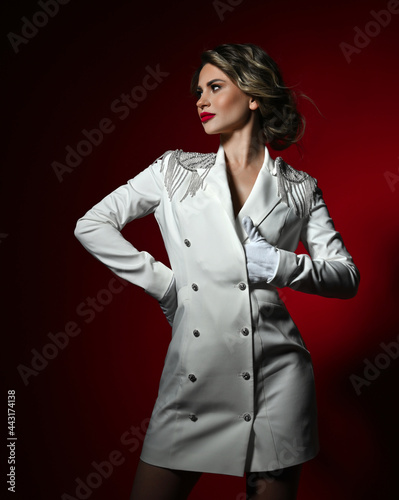 Self-confident wealthy blonde woman with permanent makeup in long white jacket with silver epaulettes and gloves stands like Napoleon army general holding, tucking hand in her bosom and looks aside