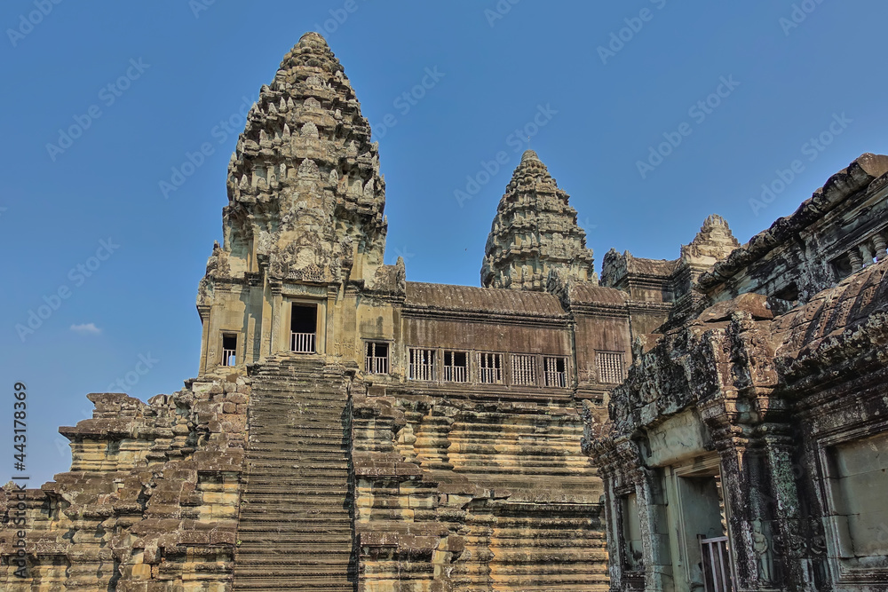 Fragment of an ancient temple in the famous Angkor. A dilapidated stone staircase leads to a terrace with a gallery. High carved towers against a clear blue sky. Cambodia