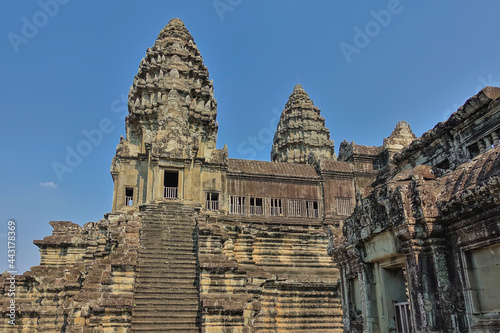 Fragment of an ancient temple in the famous Angkor. A dilapidated stone staircase leads to a terrace with a gallery. High carved towers against a clear blue sky. Cambodia