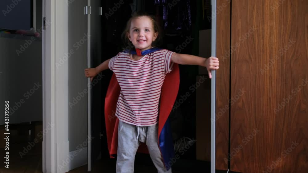 Funny preschooler girl in blue red superhero cloak laughs and appears from behind wardrobe doors with large mirrors at home Funny preschooler girl in blue red superhero cloak laughs and appears from behind wardrobe doors with large mirrors at home