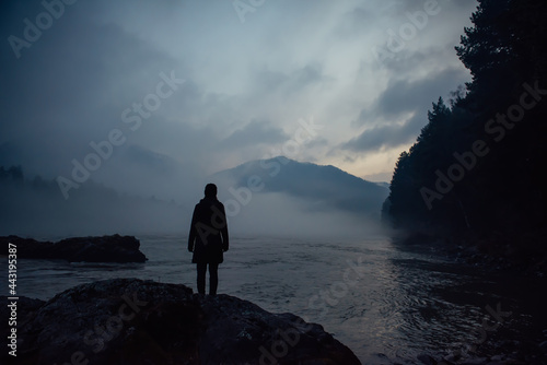 Alone human silhouette in white haze against the mountains and river. Thick fog in the evening twilight. Mysterious atmosphere. Reflection, meditation.