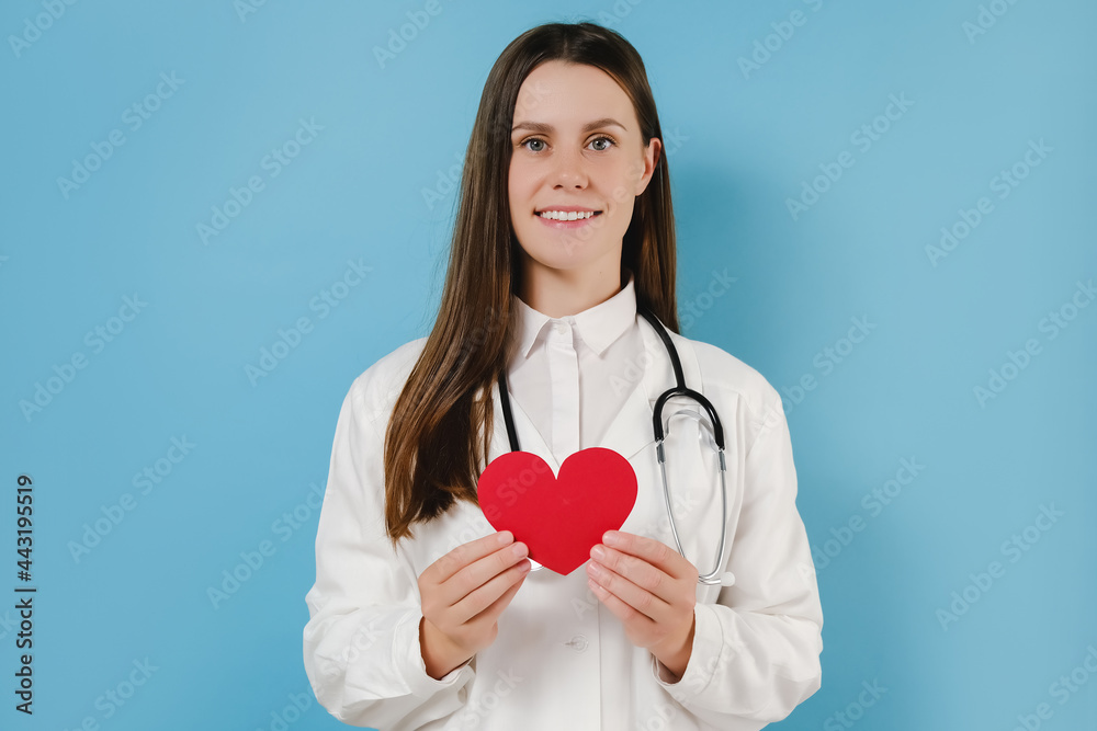 Portrait of friendly smiling young female doctor in professional medical white uniform with stethoscope holding small red heart, happy looking at camera, posing isolated over blue studio background Portrait of friendly smiling young female doctor in professional medical white uniform with stethoscope holding small red heart, happy looking at camera, posing isolated over blue studio background