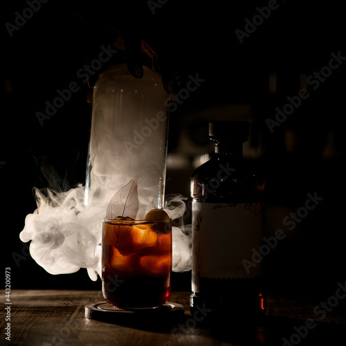 Fototapeta Close-up view of old-fashioned glass of cocktail and bottle in dark with white s